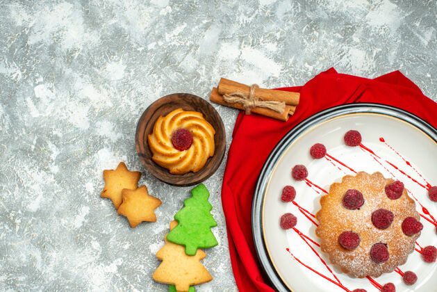顶部俯瞰白色椭圆形盘子上的浆果蛋糕红色披肩圣诞树饼干灰色表面上的自由空间早餐盘子浆果蛋糕