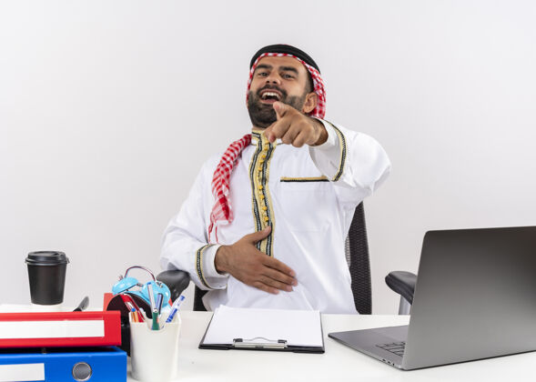 手指身着传统服装的阿拉伯商人坐在桌旁 拿着笔记本电脑笑着指着你在办公室工作电脑指坐
