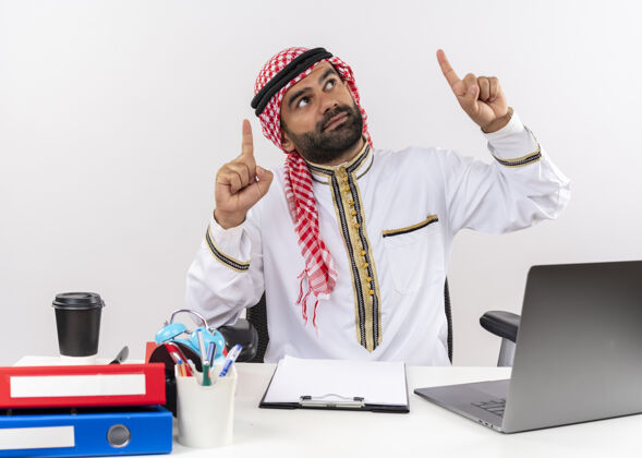 笔记本电脑身着传统服装的阿拉伯商人坐在桌旁 用食指指着笔记本电脑 看起来很自信地在办公室工作手指信心阿拉伯语