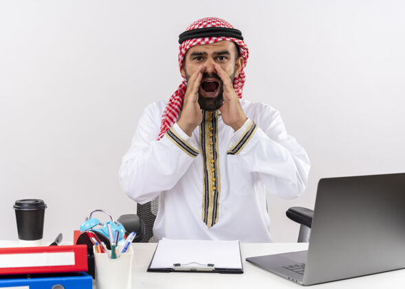 笔记本电脑身着传统服装的阿拉伯商人坐在桌旁用笔记本电脑大声叫喊 用手靠近嘴巴在办公室工作坐电脑手