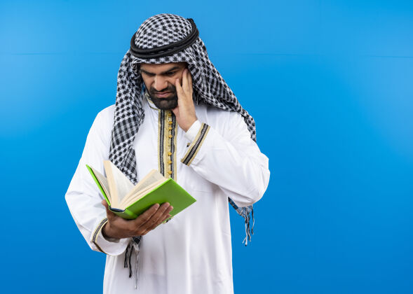 站一个穿着传统服装的阿拉伯男人站在蓝色的墙上 带着消极的情绪看着书阿拉伯语男人不高兴