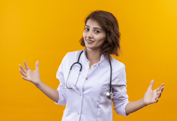 女人身穿白大褂 手持听诊器的年轻女医生做着欢迎的手势 张开双臂微笑着站在橙色的墙上医生外套手臂