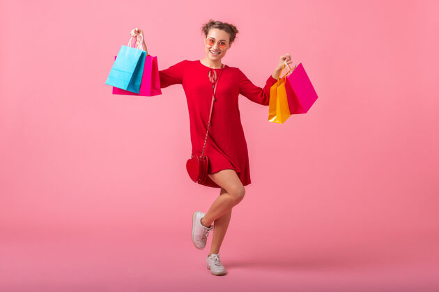快乐迷人快乐微笑的时尚女性购物狂身着红色新潮连衣裙手持五颜六色的购物袋贴在粉色的墙上 销售兴奋 春夏时尚潮流购物狂漂亮微笑