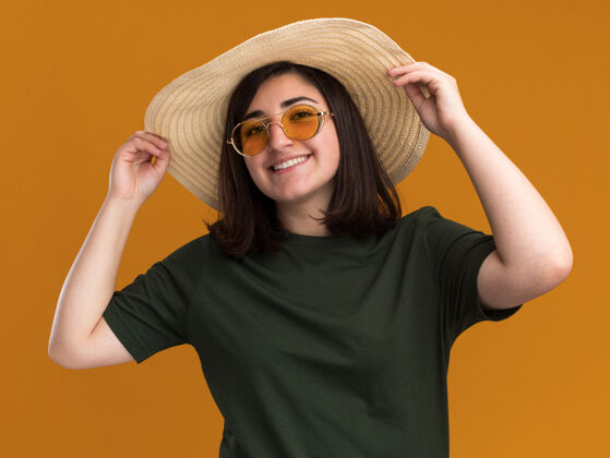 看微笑着的年轻漂亮的高加索女孩 戴着太阳眼镜 戴着沙滩帽 隔离在橙色的墙上 留着复印空间微笑太阳年轻