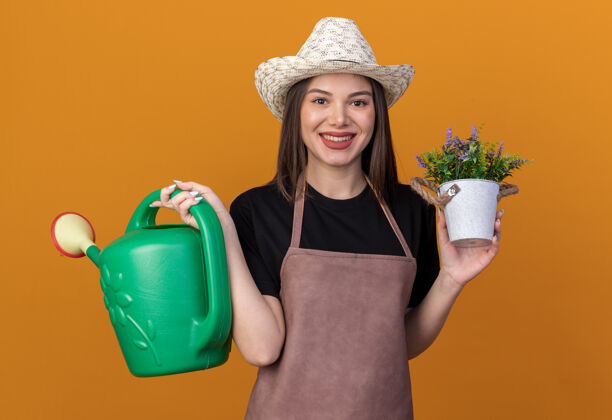 漂亮笑容可掬的白人女园丁戴着园艺帽拿着浇水罐和花盆隔离在橙色的墙上 留着复制空间园艺帽子女
