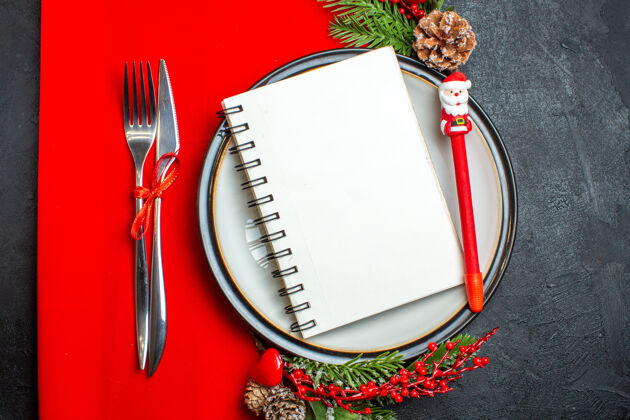 视图一个螺旋笔记本的俯视图和一支笔放在餐盘上 装饰配件杉木树枝和餐具放在红色餐巾上螺旋树枝顶部