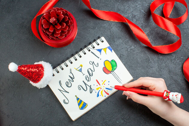 赌场手绘针叶树圆锥体的俯视图 红色丝带和笔记本 黑色背景上有新年文字和圣诞老人帽笔记本设备顶部