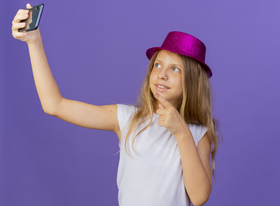 手机戴着节日帽子的漂亮小女孩用智能手机自拍微笑自信 紫色背景下的生日派对概念生日节日紫色