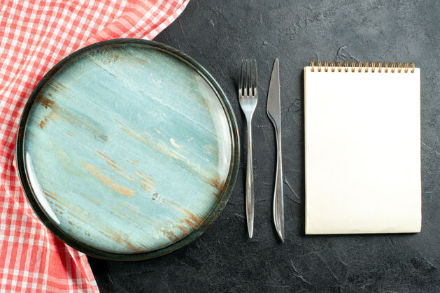 食品顶视图圆盘钢叉和餐刀红白格子桌布笔记本放在黑桌子上圆金属桌子