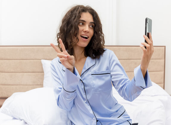 女士穿着蓝色睡衣的年轻美女坐在床上 用智能手机显示v型标志 在卧室内部灯光背景下微笑自拍卧室秀手机