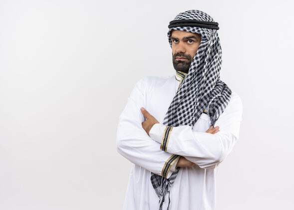 站着身着传统服装的阿拉伯男人 双臂交叉放在胸前 严肃的脸站在白色的墙壁上向一边看胸部靠边严肃