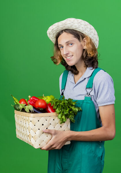蔬菜年轻的园丁穿着连体衣戴着帽子拿着装满新鲜蔬菜的箱子微笑抱着新鲜