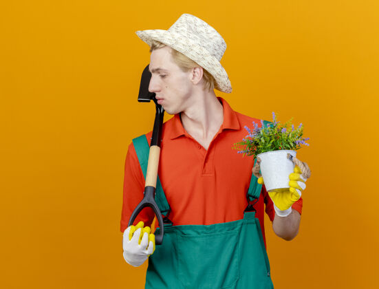 帽子年轻的园丁 穿着连体衣 戴着帽子 拿着铲子和盆栽植物 望向一边穿着抱着严肃