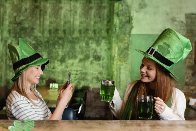 圣帕特里克日快乐的女性朋友一起庆祝圣帕特里克节与饮料和智能手机女性三叶草手机