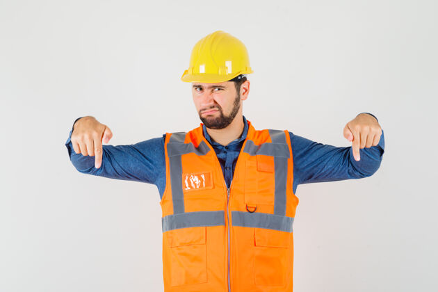 工人年轻的建筑工人穿着衬衫 背心 头盔 手指朝下 神情不满 前视建筑衬衫男人