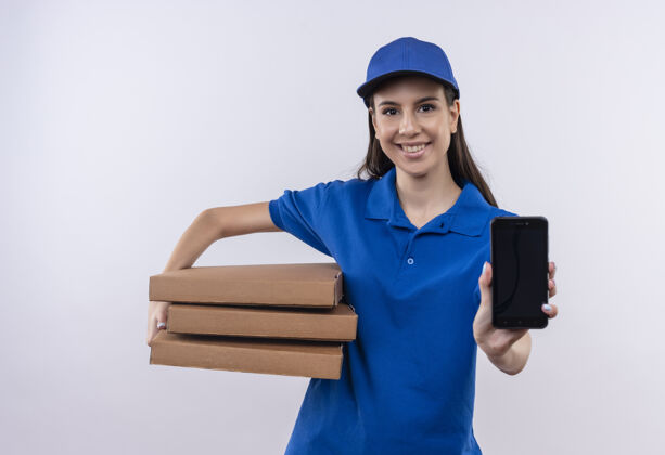 披萨身穿蓝色制服 戴着帽子的年轻送货女孩手里拿着一叠比萨饼盒 展示着智能手机看着相机 开心地微笑着送货欢呼帽子