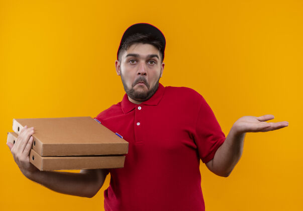 送货年轻的送货员身穿红色制服 头戴鸭舌帽 手里拿着一叠披萨盒 神情不确定 困惑不解 举起手臂没有回答制服举起看