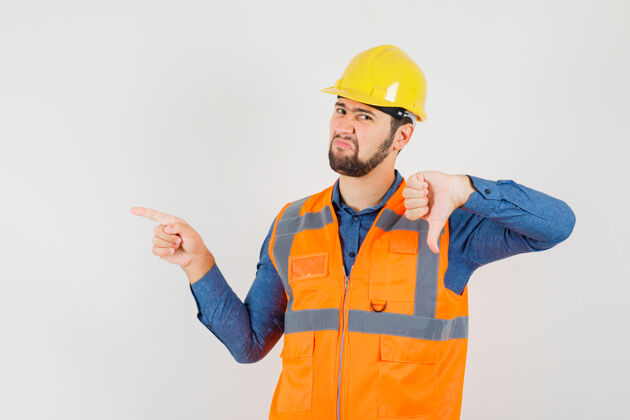 工人年轻的建筑工人穿着衬衫 背心 头盔 拇指朝下 指着侧面 神情不满 前视制服职业背心
