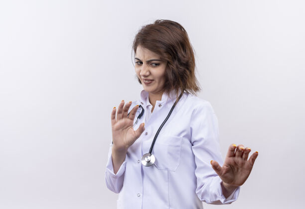厌恶身穿白大褂 手持听诊器的年轻女医生站在白墙上 用厌恶的表情做着自卫的手势年轻防御站立