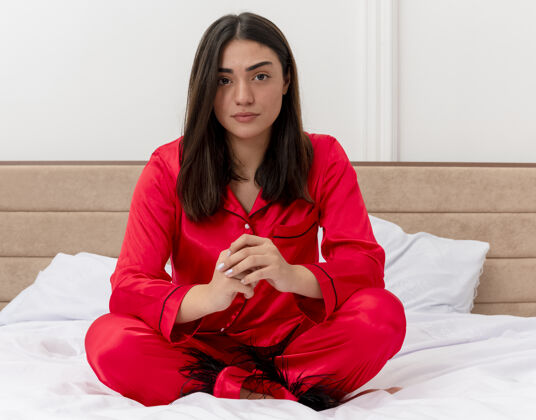 女人穿着红色睡衣的年轻漂亮女人坐在床上 在灯光背景下严肃地看着卧室内部的摄像头红色年轻看着