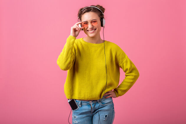 耳机迷人的快乐有趣的女人戴着耳机听音乐穿着时髦的五颜六色的服装隔离在粉红色的墙上 穿着黄色的毛衣和太阳镜 玩得很开心颜色漂亮可爱