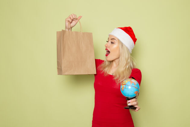 圣诞节正面图美女手持小地球仪和绿色墙上的包装圣诞彩雪节日新年感慨地球仪成人漂亮的女人