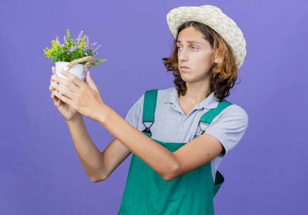 男人年轻的园丁穿着连体衣戴着帽子拿着盆栽植物罐子连身衣抱着