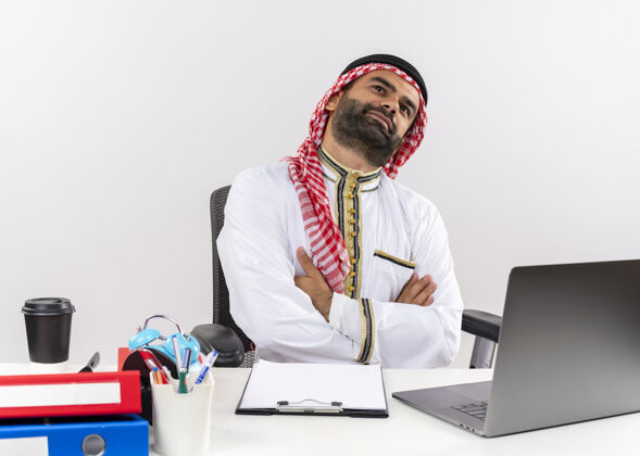 表情身着传统服装的阿拉伯商人坐在桌旁 手持笔记本电脑 面带梦幻般的表情 在办公室工作商务人士传统电脑