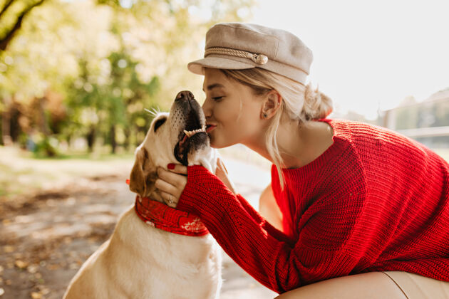 宠物美丽的年轻金发女郎穿着红色毛衣 戴着浅色帽子 在秋天的公园里与她的拉布拉多犬亲吻美丽的女孩和宠物在户外度过了完美的阳光周末多彩公园活跃