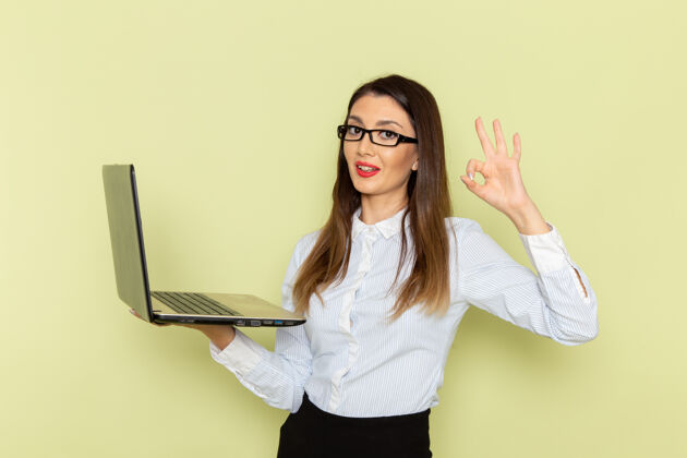 工作穿着白衬衫和黑裙子的女上班族在绿色的墙上微笑着拿着和使用她的笔记本电脑忙拿着人