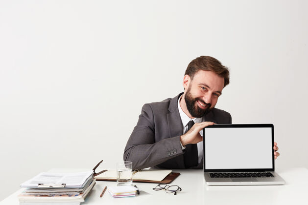 快乐年轻帅气的商人 留着棕色短发 穿着灰色西装 坐在白墙上的工作桌前 露出笔记本电脑的屏幕 笑容满面深色爱头发