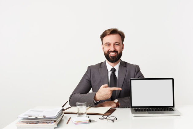 坐着照片拍摄的是一位年轻的黑发商人 留着胡子 穿着灰色西装 打着领带 拿着笔记本电脑坐在工作桌前 指着屏幕 开心地微笑着 隔着白色的墙壁男性男士正式
