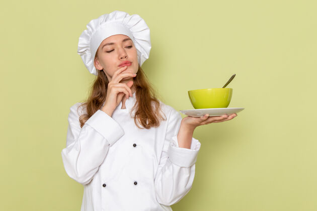 淋浴帽穿着白色厨师服的女厨师的正面图思考并在绿色墙上拿着绿色盘子女厨师烹饪制服