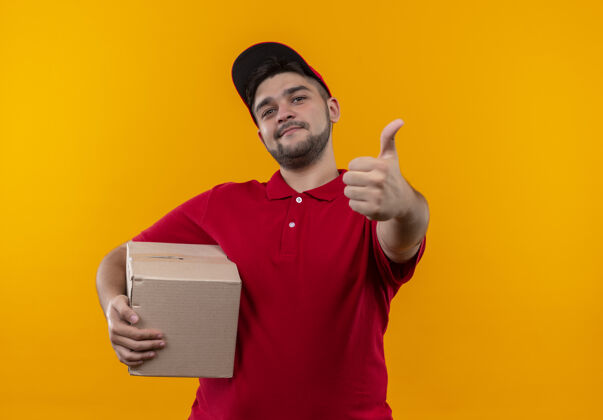 抱着年轻的送货员穿着红色制服 戴着帽子拿着盒子 微笑着自信地竖起大拇指盒子制服年轻