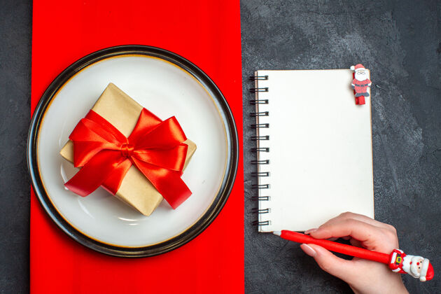 国家上图为国家圣诞大餐背景 礼物上有蝴蝶结形状的红丝带 空盘子上有红色餐巾 手拿笔放在黑色背景的笔记本上鞠躬钢笔笔记本