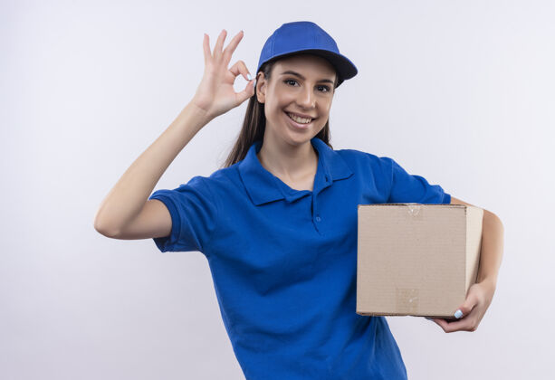 送货身着蓝色制服和帽子的年轻送货女孩抱着盒子 微笑着自信地展示着ok标志站着Ok制服