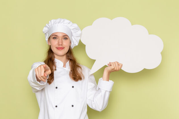 前面身着白色厨师服的女厨师正对着绿色墙上挂着微笑的白色招牌肖像西装脸