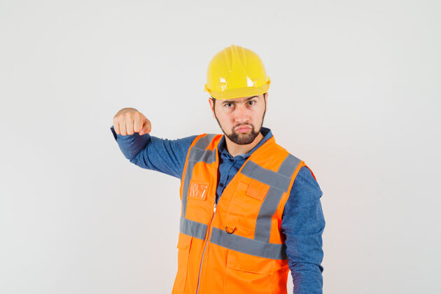 专业年轻的建筑工人穿着衬衫 背心 头盔 用拳头威胁 看起来很严肃 正面视图建筑年轻安全