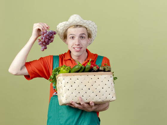 板条箱年轻的园丁穿着连体衣 戴着帽子 手里拿着装满蔬菜和葡萄的箱子 站在明亮的背景下 面带微笑地看着摄像机蔬菜束看