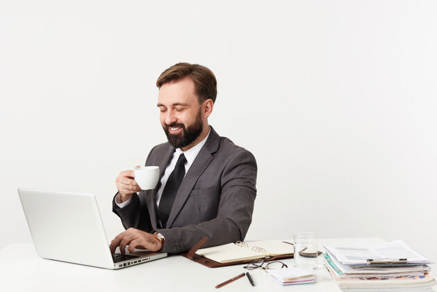 帅气工作室拍摄了一个快乐的留着胡子的黑发男人 穿着正式的衣服 拿着笔记本电脑和笔记在办公室工作 一边喝咖啡一边用手和键盘正式咖啡微笑