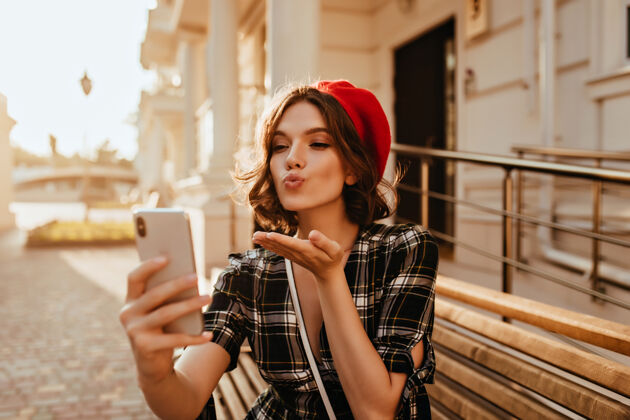 情绪迷人的黑发女人在阳光明媚的日子里一边自拍一边送上飞吻优雅的法国女模特手持智能手机拍照法国女人帽子街道