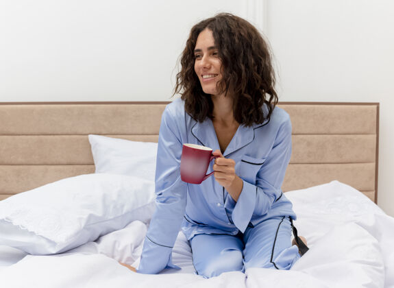 咖啡穿着蓝色睡衣的有趣年轻漂亮女人坐在床上 端着一杯咖啡 在卧室的室内灯光背景下愉快地微笑着室内床微笑