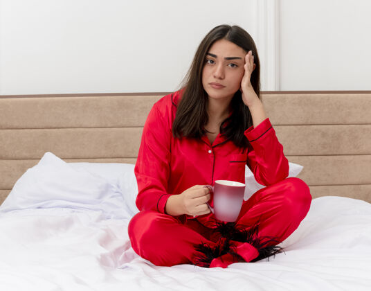 女人穿着红色睡衣的年轻美女坐在床上 端着一杯咖啡 看着摄像机 在卧室的室内灯光背景下迷茫卧室红色室内
