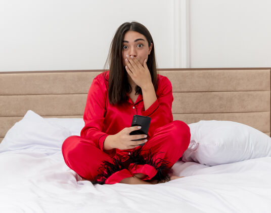 手穿着红色睡衣的年轻美女坐在床上看着摄像头 惊讶地用手捂住嘴巴 在卧室内部的灯光背景下使用智能手机漂亮卧室女人