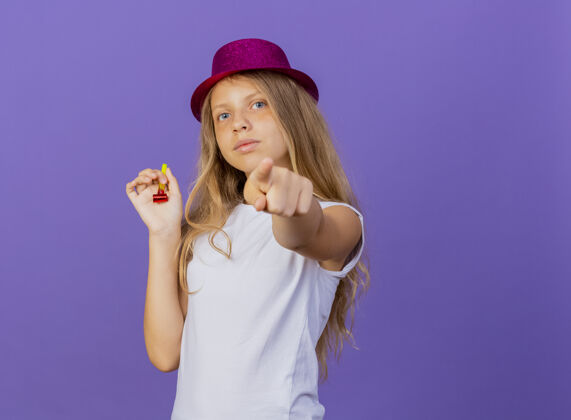 脸戴着节日礼帽的漂亮小女孩用食指指着镜头 严肃的脸 紫色背景下站着生日派对的概念相机生日派对