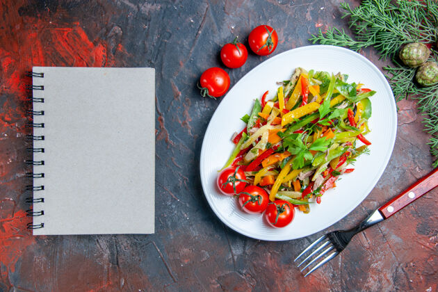 辣椒俯瞰蔬菜沙拉在椭圆形盘子叉樱桃西红柿笔记本在深红色的桌子上辣椒膳食辣椒