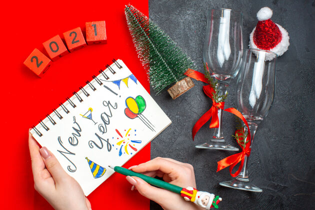 高脚杯手持螺旋笔记本的顶视图 黑色和红色背景上有新年图画和圣诞树玻璃高脚杯数字玻璃设备圣诞节