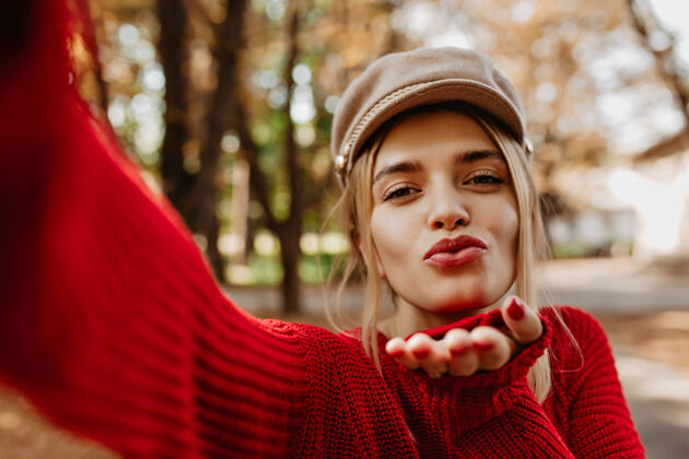 户外美丽的金发女孩在公园里自拍时亲吻可爱的女人穿着时髦的红色毛衣和轻便的帽子日落自拍乡村