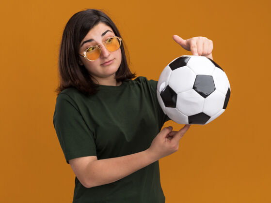 球印象深刻的年轻漂亮的白人女孩在太阳眼镜举行 并看着球孤立在橙色墙上的复制空间看起来眼镜抱着