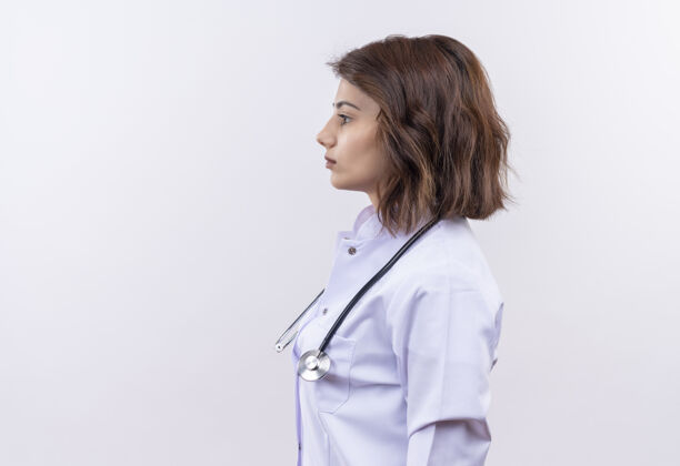 严肃身穿白大褂 手持听诊器的年轻女医生站在一边 严肃的脸覆盖着白色的背景年轻站着外套